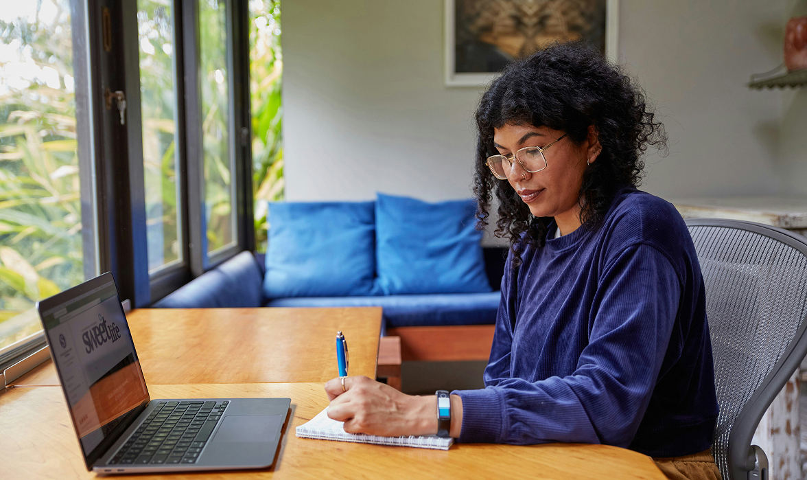Professional taking notes while working on a laptop in a bright home office, representing remote work, productivity, and online collaboration.