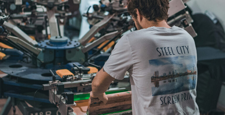Worker operating a screen printing press to produce custom printed T-shirts in a commercial print shop.